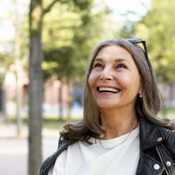 Cheerful overjoyed middle aged woman wearing sunglasses on her head and black leather jacket over white blouse enjoying peaceful beautiful morning while waking in park, looking up with broad smile